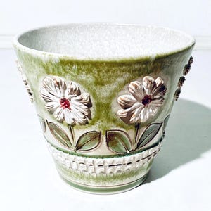 Könnte beinhalten: Ein Blumentopf aus Keramik mit grüner und weißer Glasur. Der Topf hat geprägte weiße Blumen mit roten Zentren und grünen Blättern. Der Boden hat ein strukturiertes Korbgeflecht-Design. Ein dekoratives Stück für Pflanzen.