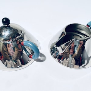 Könnte beinhalten: Ein silbernes Zucker- und Milchkännchen-Set mit blauen und roten Griffen. Die Zuckerdose hat einen schwarzen Knopf auf dem Deckel. Beide Teile haben eine glänzende, reflektierende Oberfläche.