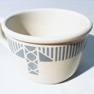 Könnte beinhalten: Eine weiße Keramiktasse mit einem grauen geometrischen Design. Das Design zeigt Streifen, Kreise und Dreiecke.