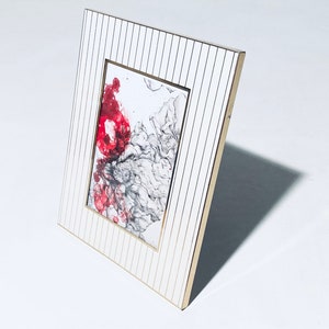 Könnte beinhalten: Weißer Bilderrahmen mit goldener Verzierung und vertikalen Linien. Der Rahmen enthält ein Gemälde mit abstrakter Kunst in Rot und Schwarz.