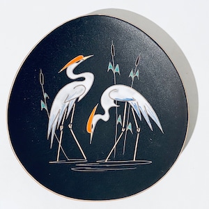 May include: A black ceramic plate with a white and blue design of two birds standing in a marsh. The birds have orange beaks and are surrounded by green reeds.