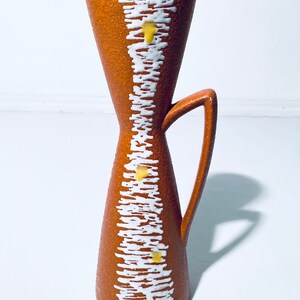 Puede incluir: Un jarr&oacute;n de cer&aacute;mica marr&oacute;n con un dise&ntilde;o geom&eacute;trico blanco y amarillo. El jarr&oacute;n tiene un asa y una abertura estrecha.