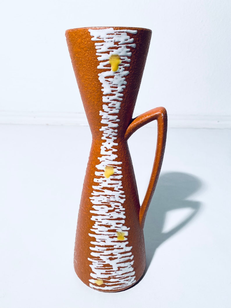 Puede incluir: Un jarr&oacute;n de cer&aacute;mica marr&oacute;n con un dise&ntilde;o geom&eacute;trico blanco y amarillo. El jarr&oacute;n tiene un asa y una abertura estrecha.