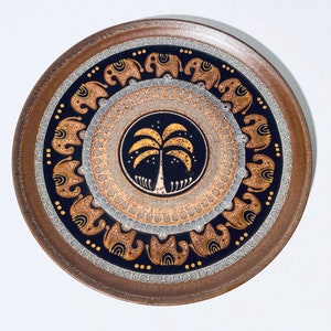 May include: Round brown ceramic plate with a decorative design. The plate features a central palm tree motif in gold and white, surrounded by concentric circles with elephant and dot patterns. The rim is a darker brown.