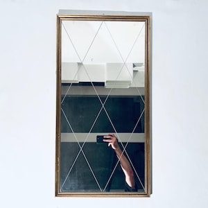 Könnte beinhalten: Rechteckiger Spiegel mit goldenem Rahmen und Rautenmuster auf dem Glas. Der Spiegel reflektiert einen Raum und eine Person, die ein Telefon hält. Er hängt an einer weißen Wand.