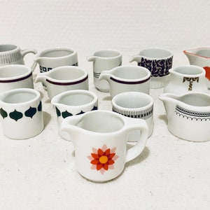 May include: A collection of 11 small white ceramic creamer pitchers with various black, brown, and red designs. The pitchers have a variety of patterns, including geometric shapes, flowers, and hearts.