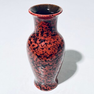 Könnte beinhalten: Eine kleine rote und schwarze, gesprenkelte Keramikvase. Die Vase hat einen abgerundeten Boden und eine schmale Öffnung.