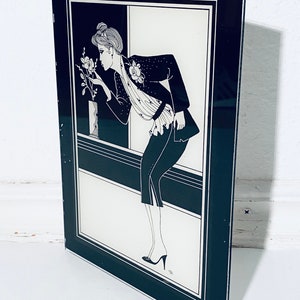 May include: Black and white framed illustration of a woman in a black jacket and skirt, looking at a flower in a vase. The illustration is set against a white background.