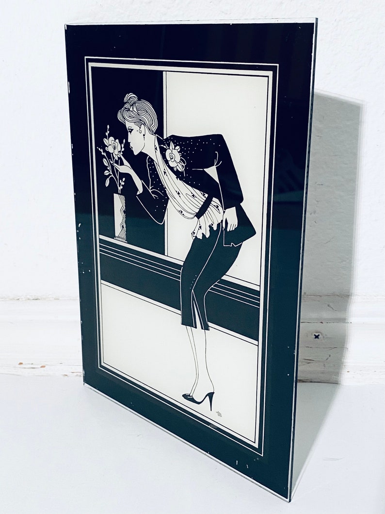 May include: Black and white framed illustration of a woman in a black jacket and skirt, looking at a flower in a vase. The illustration is set against a white background.