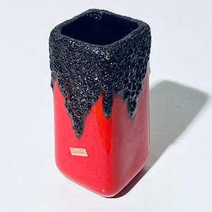 Puede incluir: Un jarrón de cerámica rojo y negro con un esmalte texturizado, similar a la lava. El jarrón tiene forma cuadrada y tiene una pequeña etiqueta en el lateral que dice "Alemania Occidental".