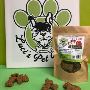 May include: A brown paper bag with a white label that says "Disobedient Flavors! The BCR! Guilt Free Small Batch Dog Treats Bacon Chicken Ranch 100% USA Sourced Ingredients" and a picture of a dog. There are four brown dog treats on a green surface.
