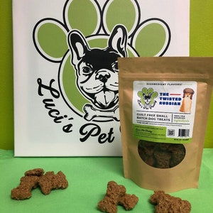 May include: A brown paper bag with a white label that says "The Twisted Russian" and "Guilt Free Small Batch Dog Treats". The bag is filled with small, brown dog treats. The bag is sitting on a green surface with a few dog treats scattered around it.