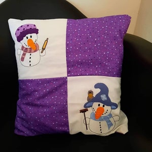 May include: A purple and white cushion with two embroidered snowmen. One snowman is holding a rolling pin and the other is holding a broom. The background of the cushion is a purple fabric with white hearts.