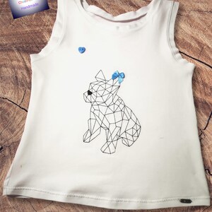 以下が含まれることがあります： 幾何学模様の犬のデザインが施された白いノースリーブシャツ。犬は黒で縁取られ、青いリボンが付いています。青いハート型のボタンがシャツに縫い付けられています。「Bine's Chaos-Ecke Handmade」のブランド名が左上に表示されています。