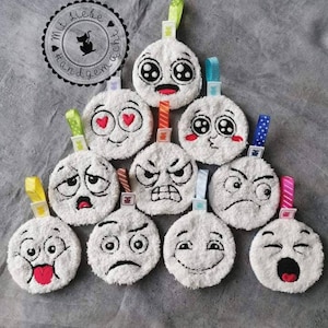 Peut inclure: Dix cercles ronds en tissu pelucheux blanc avec des expressions faciales brodées. Chaque cercle est doté d'un ruban coloré. Les cercles sont disposés en forme de pyramide. Le texte "Mit Liebe Handgemacht" est sur un cercle noir avec une silhouette de chat blanc.