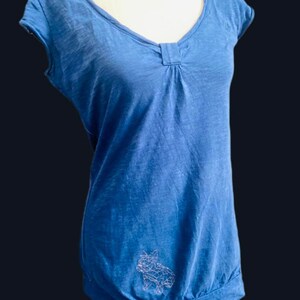 以下が含まれることがあります： Vネックとネックラインにギャザーディテールが付いた青い半袖トップス。裾はギャザー。白い幾何学的な犬のデザインがシャツの左下に刺繍されています。