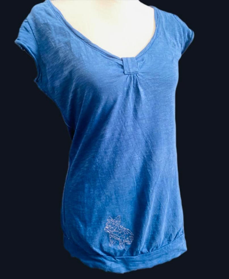 以下が含まれることがあります： Vネックとネックラインにギャザーディテールが付いた青い半袖トップス。裾はギャザー。白い幾何学的な犬のデザインがシャツの左下に刺繍されています。