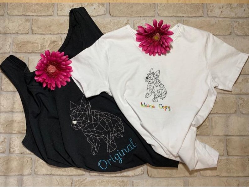 以下が含まれることがあります： 幾何学的な犬のデザインが施された黒と白の2枚のTシャツ。黒いシャツには青で「Original」の文字、白いシャツには虹色で「Meine Copy」の文字が書かれています。ピンクの花がシャツを飾っています。