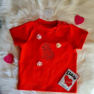 以下が含まれることがあります： 幾何学的な犬のデザインが施された、鮮やかな赤色のベビーTシャツ。シャツには小さな花のアクセントと、「Elara」と2つのハートが描かれたパッチが付いています。Tシャツはふわふわの白い表面に置かれています。