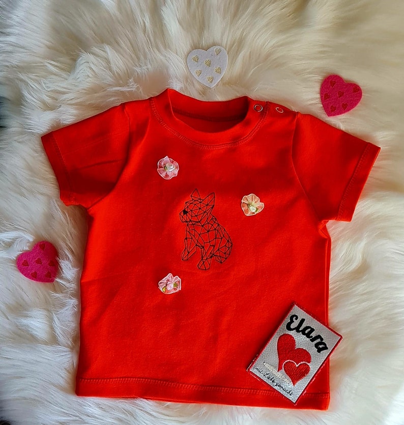 以下が含まれることがあります： 幾何学的な犬のデザインが施された、鮮やかな赤色のベビーTシャツ。シャツには小さな花のアクセントと、「Elara」と2つのハートが描かれたパッチが付いています。Tシャツはふわふわの白い表面に置かれています。