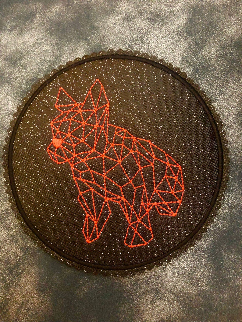 以下が含まれることがあります： オレンジ色の糸で刺繍された幾何学的なキツネのデザインが施された、丸い黒いパッチ。パッチはキラキラした背景とスカラップエッジを持っています。キツネは相互接続された線でできています。