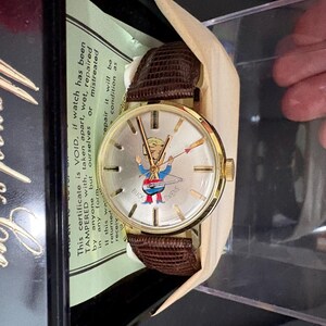 May include: A vintage gold-tone Buck Owens wristwatch with a brown leather strap. The watch face displays a cartoon figure and the words "Buck Owens". The watch is presented in its original box with a warranty certificate.