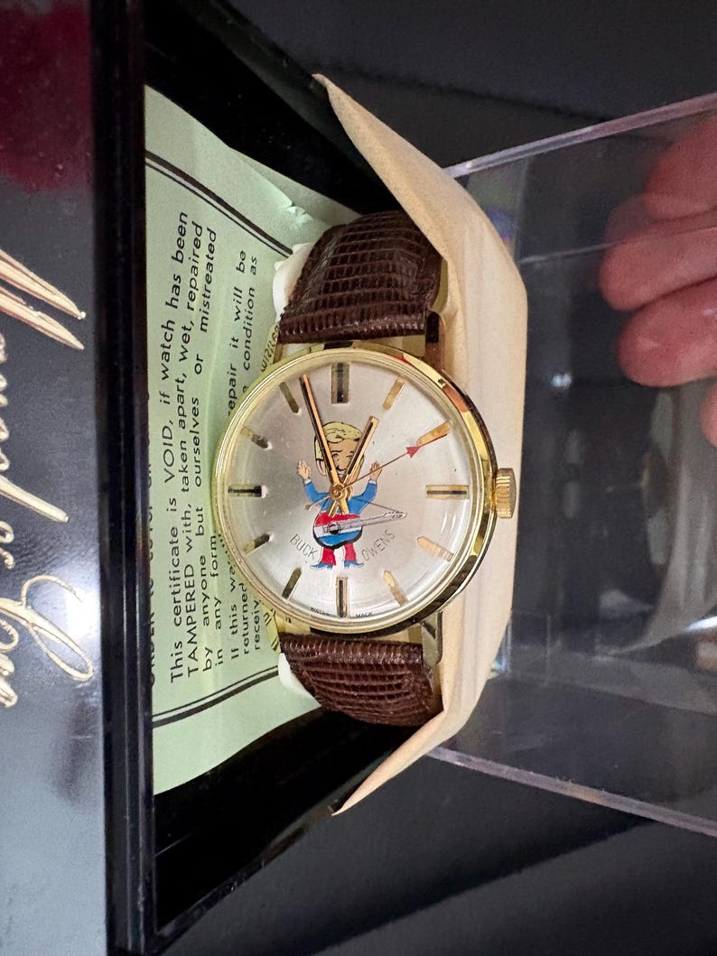 May include: A vintage gold-tone Buck Owens wristwatch with a brown leather strap. The watch face displays a cartoon figure and the words "Buck Owens". The watch is presented in its original box with a warranty certificate.