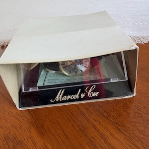 May include: A vintage watch in its original box. The watch face is visible through a clear plastic window. The box is white with the words "Marcel & Cie" in gold script on a black base.