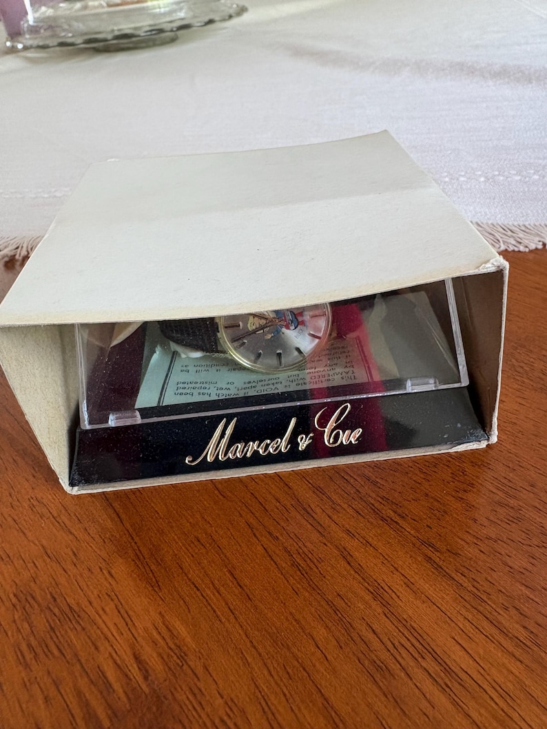 May include: A vintage watch in its original box. The watch face is visible through a clear plastic window. The box is white with the words "Marcel & Cie" in gold script on a black base.
