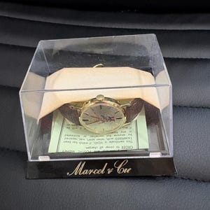 May include: A vintage gold-coloured wristwatch with a brown leather strap, presented in a clear plastic case. The watch face has a unique design. The case sits on a black base with the text "Marcel & Cie" in gold.