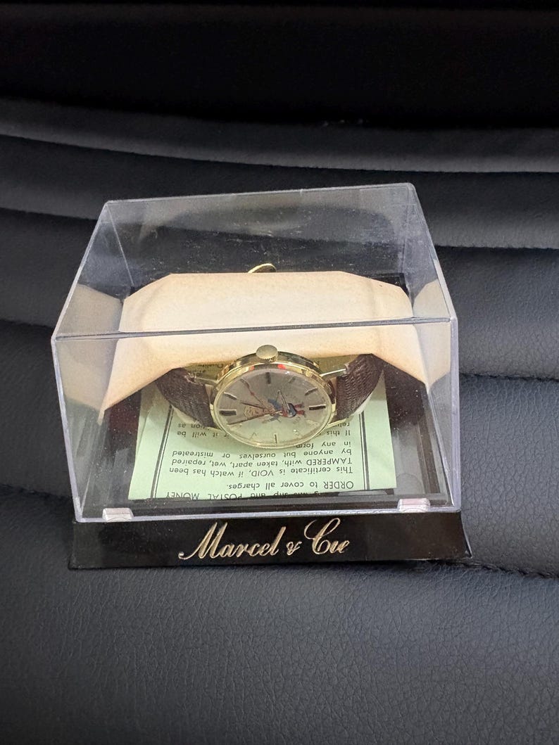 May include: A vintage gold-coloured wristwatch with a brown leather strap, presented in a clear plastic case. The watch face has a unique design. The case sits on a black base with the text "Marcel & Cie" in gold.