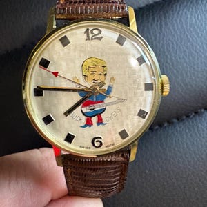 May include: A vintage gold-toned wristwatch with a brown leather strap. The watch face displays a cartoon of a man playing guitar, with "BUCK OWENS" printed on it. It has black square hour markers and a gold crown. The watch is Swiss made.