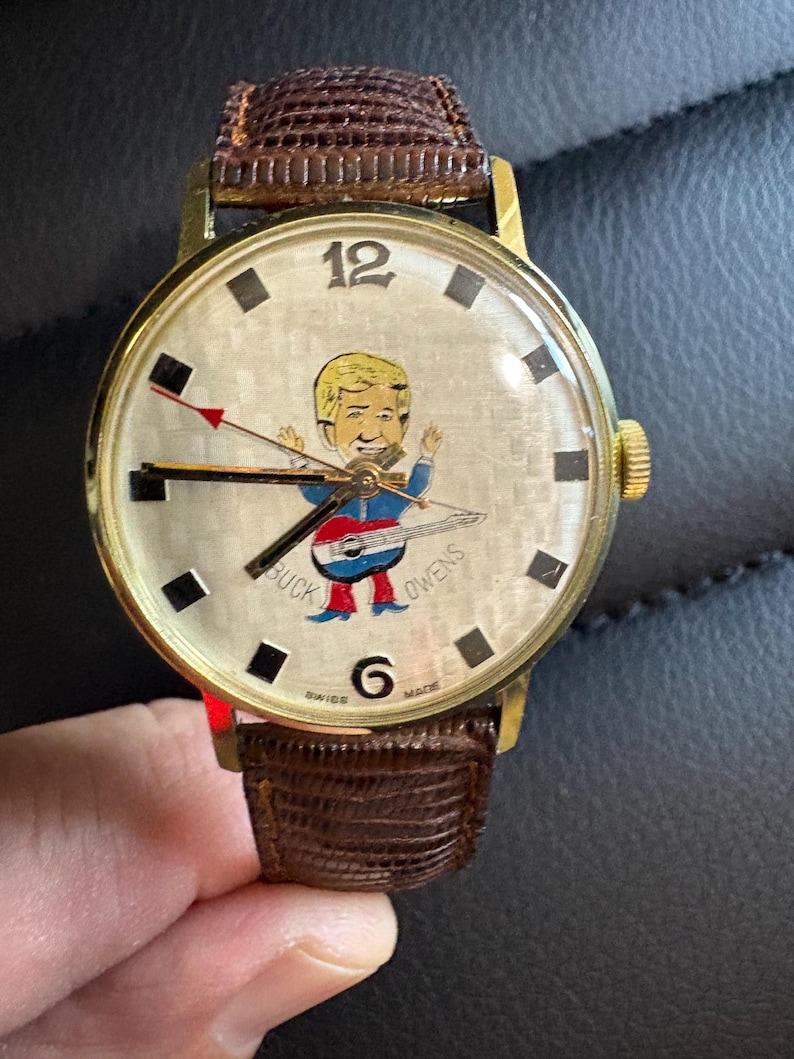 May include: A vintage gold-toned wristwatch with a brown leather strap. The watch face displays a cartoon of a man playing guitar, with "BUCK OWENS" printed on it. It has black square hour markers and a gold crown. The watch is Swiss made.