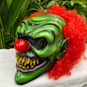 Puede incluir: Una máscara de payaso verde y roja con una gran sonrisa de dientes y una nariz roja. La máscara tiene una visera negra y está hecha de un material plástico duro. La máscara está decorada con pelo rojo.