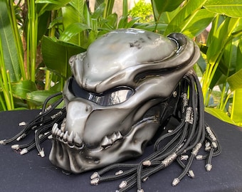 Op de afbeelding: Een zilveren motorhelm met een Predator-ontwerp. De helm heeft een zwart vizier en een zwart en zilveren gevlochten koord dat aan de achterkant is bevestigd.