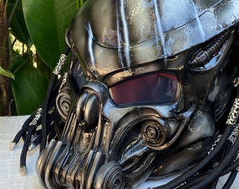 May include: A detailed, metallic Predator helmet with a glossy black and silver finish. The helmet features a red visor, intricate mechanical details, and black dreadlocks with silver accents. The design is inspired by the Predator movie franchise.