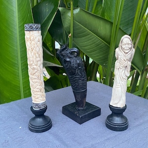Peut inclure: Trois sculptures décoratives sur socles noirs. À gauche, un pilier cylindrique crème avec un entrelacs celtique complexe. Au centre, une sculpture noire en forme de corne avec des détails texturés. À droite, une figurine crème représentant une personne âgée.