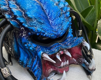 Könnte beinhalten: Blau-schwarzer Motorradhelm im Predator-Design. Der Helm hat ein strukturiertes, schuppiges Design mit scharfen Zähnen, einem bedrohlichen offenen Mund und schwarzen Dreadlocks. Das Innere des Mundes ist rot.