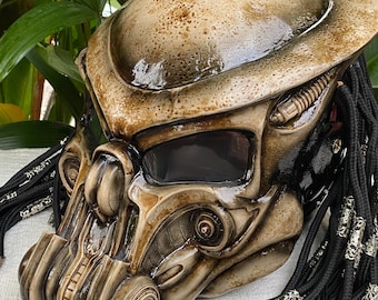 May include: A brown and black Predator-style motorcycle helmet with a black and silver braided dreadlock-style accessory. The helmet has a dark tinted visor and a detailed design.