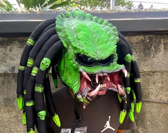 Op de afbeelding: Groene en zwarte motorhelm met een Predator-ontwerp. De helm heeft een grote, getande mond en een groen, getextureerd oppervlak. De helm is versierd met zwarte dreadlocks met groene accenten en schedelvormige bedels.