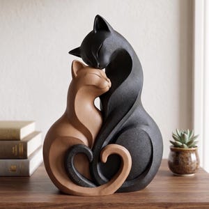 May include: A decorative sculpture featuring two stylized cats sitting together. One cat is matte black and the other is a warm tan color. Their bodies are curved and their tails intertwine to form a heart shape at the base. The cats have closed eyes.