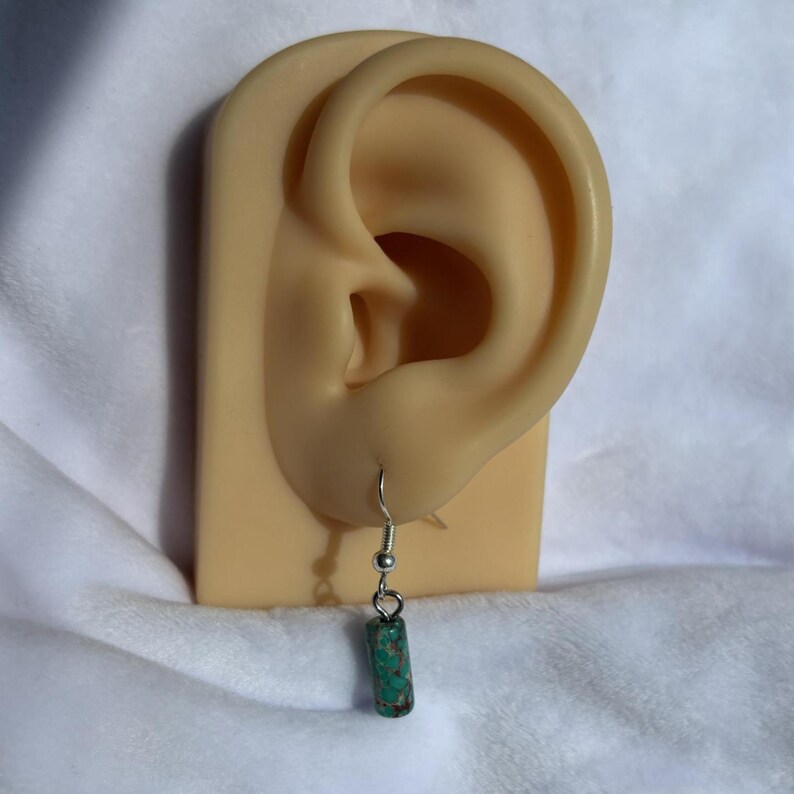 Pode incluir: Um &uacute;nico brinco com um gancho prateado e uma conta cil&iacute;ndrica de pedra turquesa e castanha. O brinco &eacute; exibido em um modelo de orelha bege, contra um fundo branco texturizado. Uma joia simples e elegante.