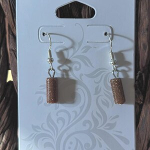 Pode incluir: Brincos pendurados com contas cil&iacute;ndricas castanhas e brilhantes. Os brincos pendem de ganchos de orelha prateados. S&atilde;o exibidos em um cart&atilde;o branco com um padr&atilde;o floral decorativo.