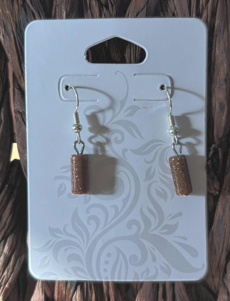 Pode incluir: Brincos pendurados com contas cil&iacute;ndricas castanhas e brilhantes. Os brincos pendem de ganchos de orelha prateados. S&atilde;o exibidos em um cart&atilde;o branco com um padr&atilde;o floral decorativo.