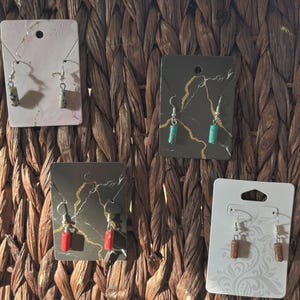 May include: A collection of handmade earrings displayed on decorative cards. The earrings feature various stone beads in shades of gray, green, red, and brown. Each pair is suspended from silver-colored ear wires, showcasing a minimalist design.