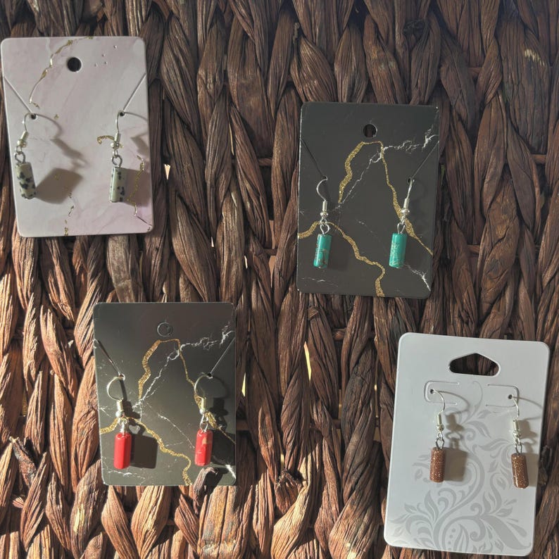 Pode incluir: Uma cole&ccedil;&atilde;o de brincos artesanais exibidos em cart&otilde;es decorativos. Os brincos apresentam contas de pedra em tons de cinza, verde, vermelho e castanho. Cada par &eacute; suspenso por ganchos prateados, mostrando um design minimalista.
