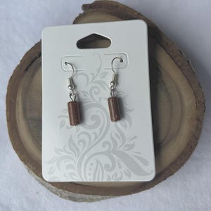 Pode incluir: Um par de brincos pendurados com contas cil&iacute;ndricas de pedra castanho-dourado. Os brincos penduram-se de ganchos de orelha prateados. S&atilde;o exibidos num cart&atilde;o branco com um padr&atilde;o decorativo.