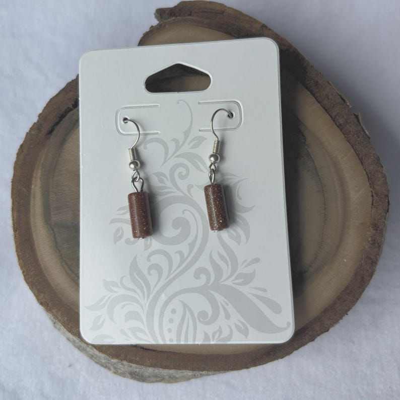 Pode incluir: Um par de brincos pendurados com contas cil&iacute;ndricas de pedra castanho-dourado. Os brincos penduram-se de ganchos de orelha prateados. S&atilde;o exibidos num cart&atilde;o branco com um padr&atilde;o decorativo.