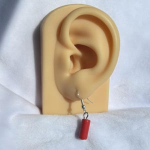 Pode incluir: Um &uacute;nico brinco cil&iacute;ndrico vermelho com um gancho prateado &eacute; exibido. O brinco est&aacute; pendurado na orelha de um manequim. O fundo &eacute; um tecido branco macio. O brinco tem um design simples e minimalista.