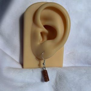 Pode incluir: Um &uacute;nico brinco com um gancho prateado e uma conta cil&iacute;ndrica marrom e brilhante. O brinco &eacute; exibido em uma forma de orelha bege contra um fundo branco texturizado.